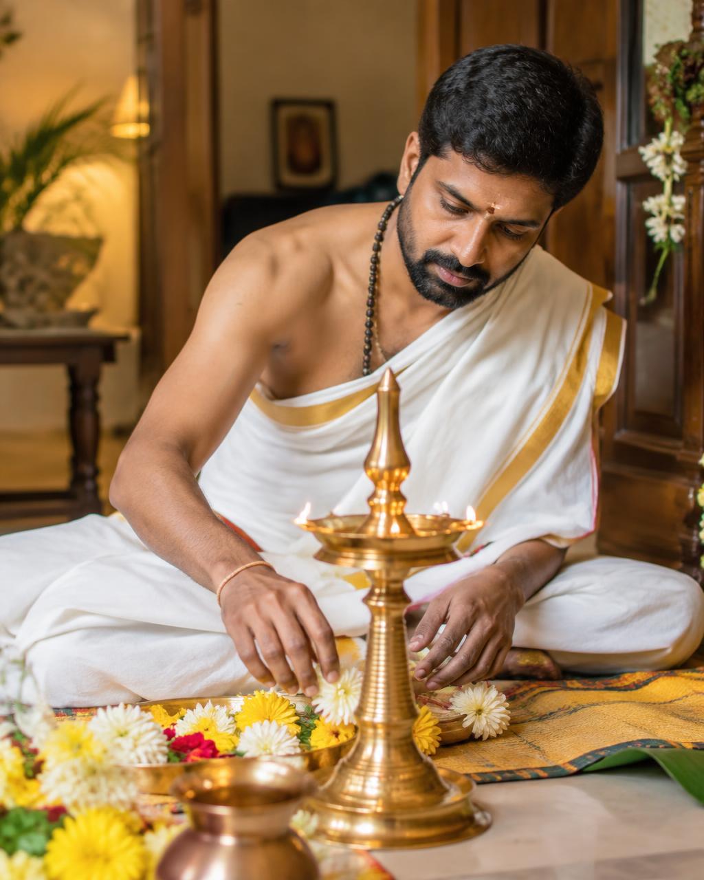 A pujari in traditional white attire arranging flowers beside a brass lamp